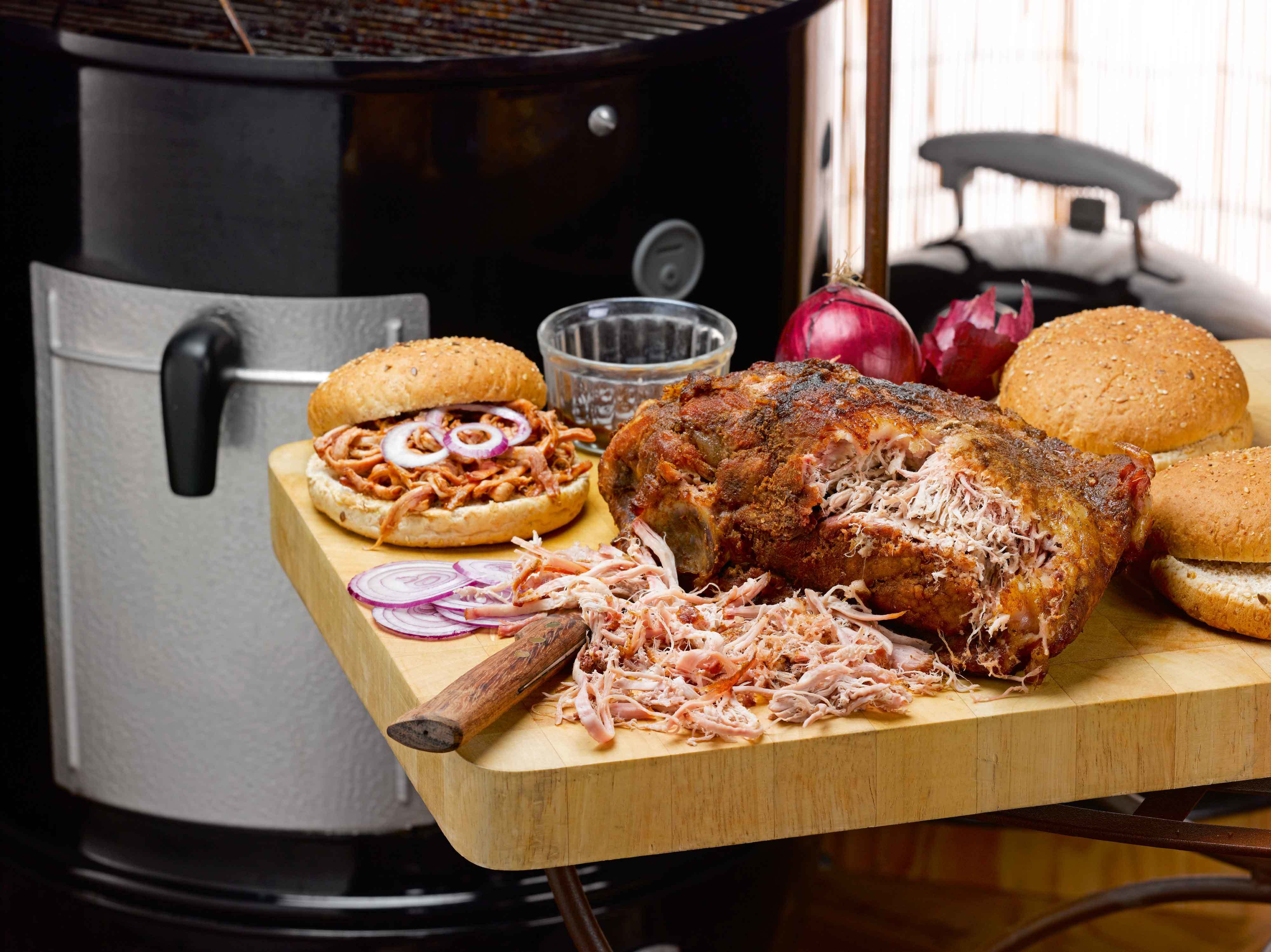 Een tafel met daarop een geroosterde varkensschouder met een deel eruit gesneden om pulled pork van te maken met een broodje pulled pork ernaast